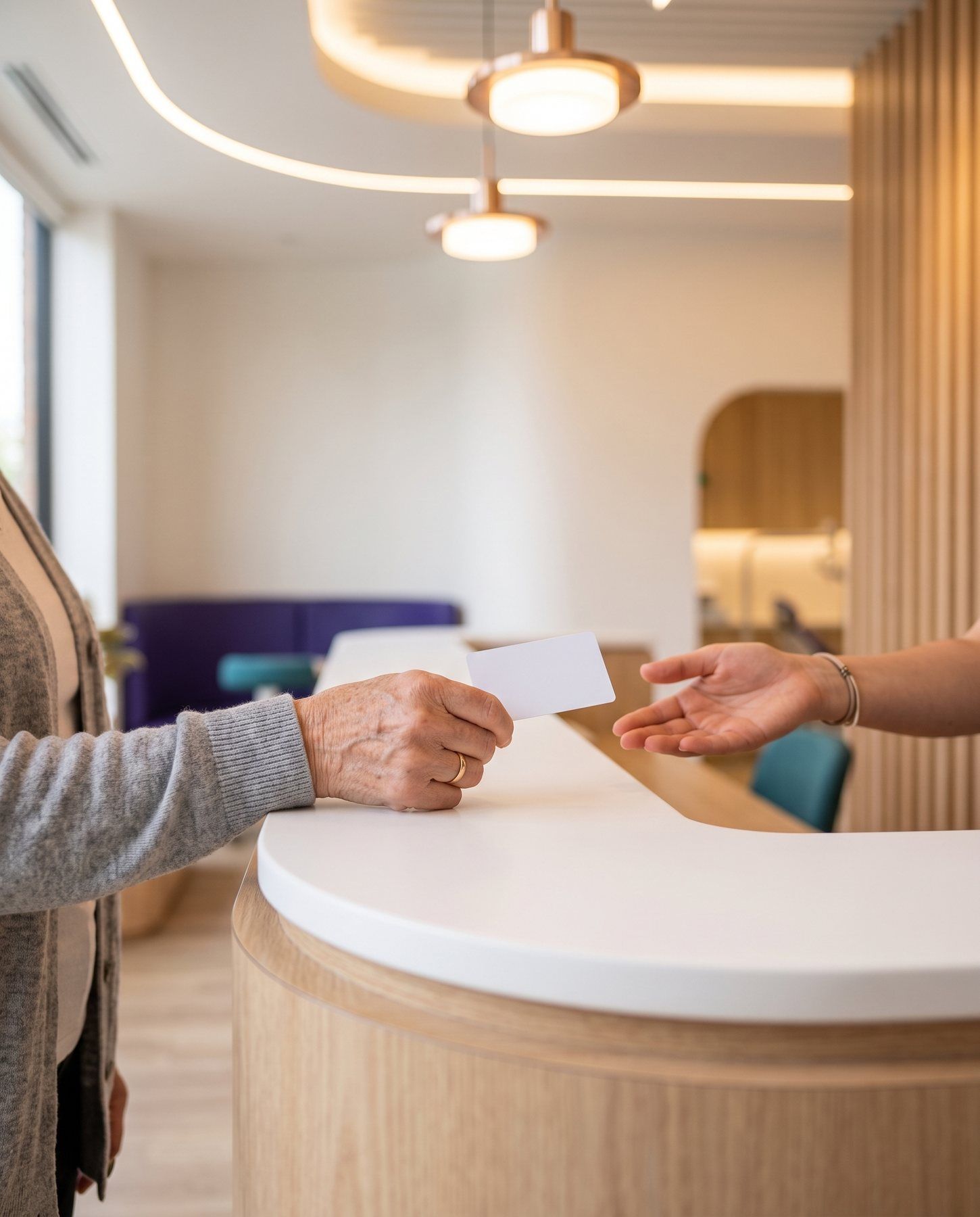 An older patient's hand passing a card across a light oak reception counter to a receptionist